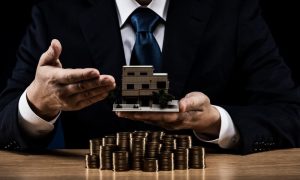 Man holding a house model near a pile of coins while thinking about rental property passive income