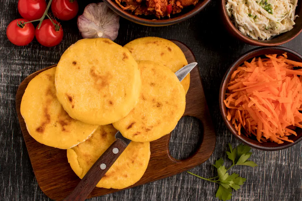 A traditional Salvadoran dish of pupusas stacked on a wooden board, surrounded by sides of shredded carrots, cabbage slaw, tomatoes, garlic, and other garnishes.