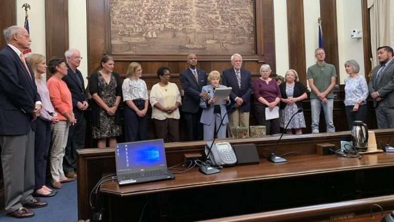 Formal ceremony in wood-paneled government chamber with group of officials and citizens gathered around podium, American flag visible, and historical Alexandria map on wall