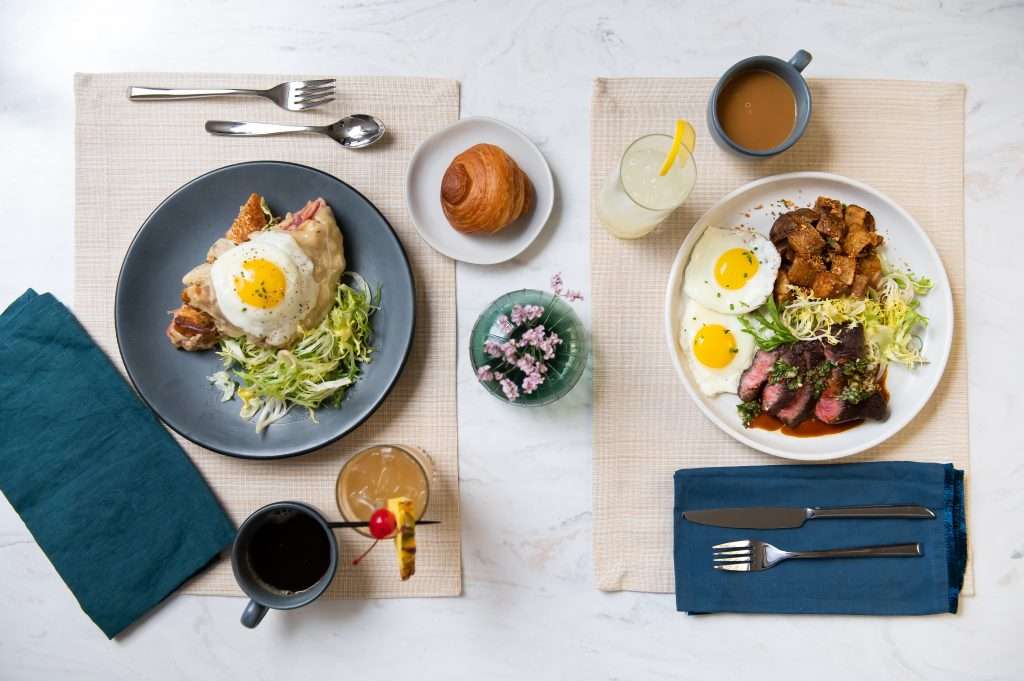 Meals at Ted's Bulletin feature steak and eggs, croque madame, croissant, drinks, and coffee on placemats with cutlery.