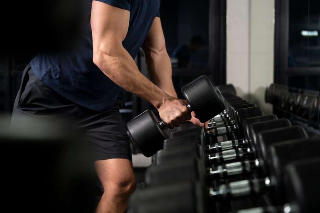 Man in navy athletic wear selecting heavy dumbbells from weight rack in modern gym with professional lighting showing muscular arm definition