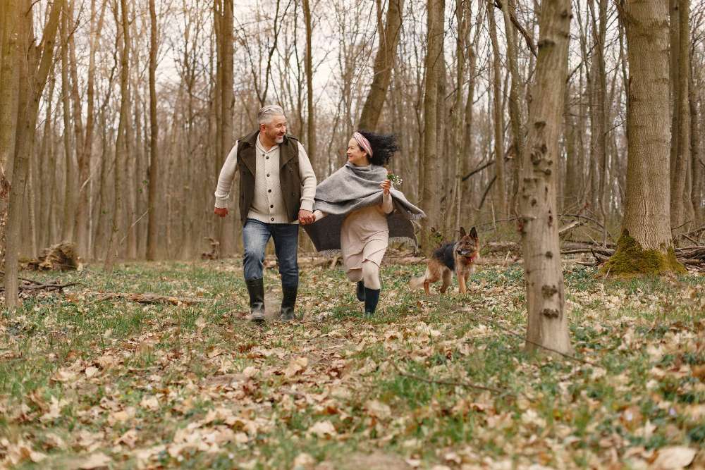 Elderly couple joyfully walking hand in hand through a forest, accompanied by a German Shepherd.