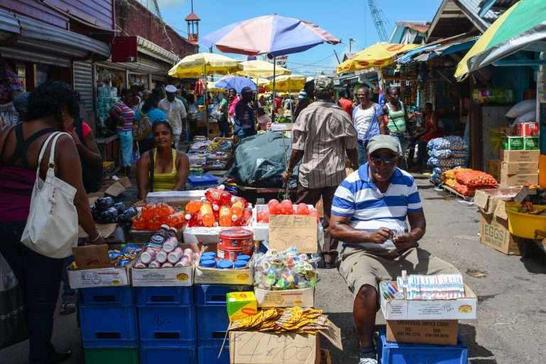 A lively outdoor market scene in Georgetown, Guyana, with vendors selling colorful goods like beverages, snacks, and produce under bright umbrellas, while shoppers and pedestrians fill the crowded walkway on a sunny day.