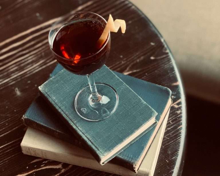 Captain Gregory's elegant cocktail service featuring dark amber drink with lemon twist garnish in wine glass placed on vintage blue books on wooden table with dramatic lighting
