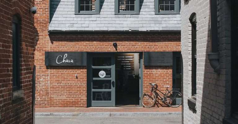 A brick building with a gray door and slate roof housing Chaia Tacos, with a black sign above the entrance and a bicycle parked outside along the sidewalk.