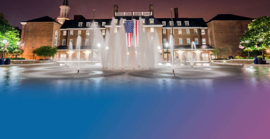 City of Alexandria Government building at night with dramatic water fountains illuminated in blue and purple lights, large American flag displayed on historic brick facade