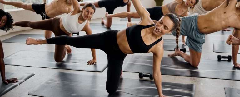 Group fitness class performing side plank exercises with extended legs on yoga mats in bright CorePower Yoga studio with natural lighting