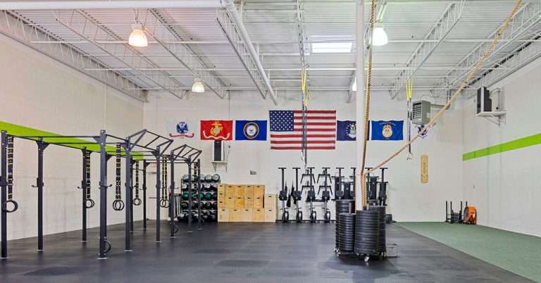 Spacious CrossFit gym featuring pull-up rigs, gymnastic rings, ropes, medicine balls, plyometric boxes, and rowing machines, with American and military flags on the wall.
