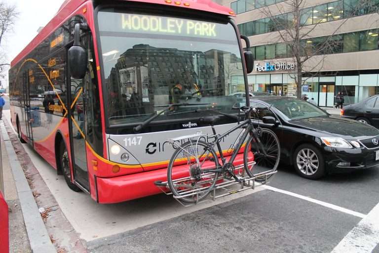 Red DC Circulator bus on city street near FedEx Office with bicycle mounted on front rack traveling toward Woodley Park in Washington DC