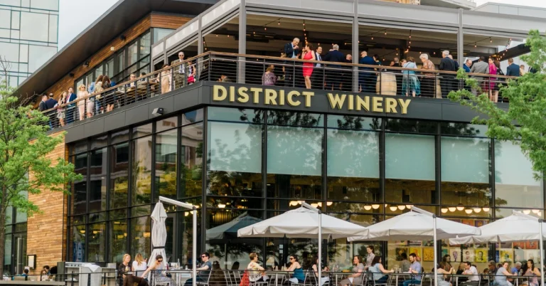Crowd gathered on the upper deck of District Winery in Washington, D.C., with glass-paneled architecture and outdoor patio seating below.