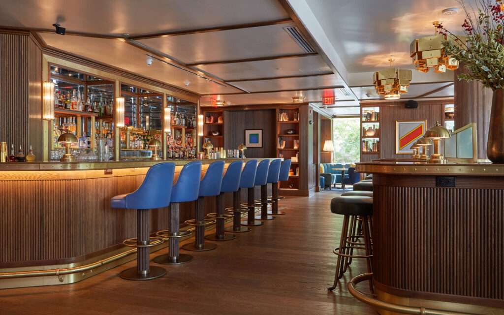 Doyle Bar elegant interior featuring rich wood paneling, brass accents, blue leather bar stools, illuminated bar display, and sophisticated mid-century modern design elements