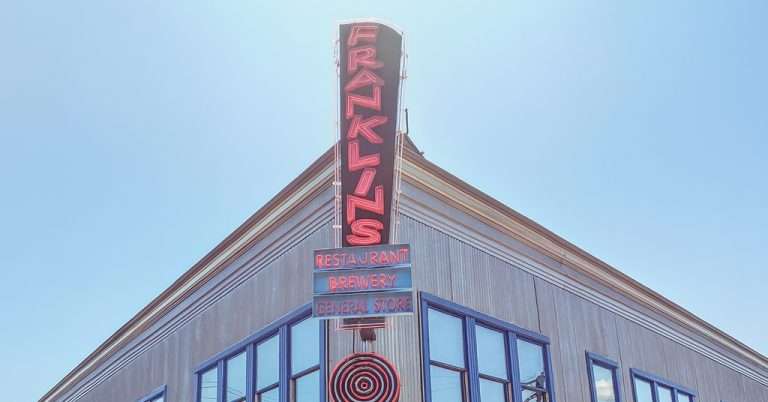 A corner view of Franklin’s Brewery, featuring a large vertical neon sign reading “Franklin’s” with “Restaurant,” “Brewery,” and “General Store” beneath, mounted on a rustic building with blue-trimmed windows against a clear blue sky.