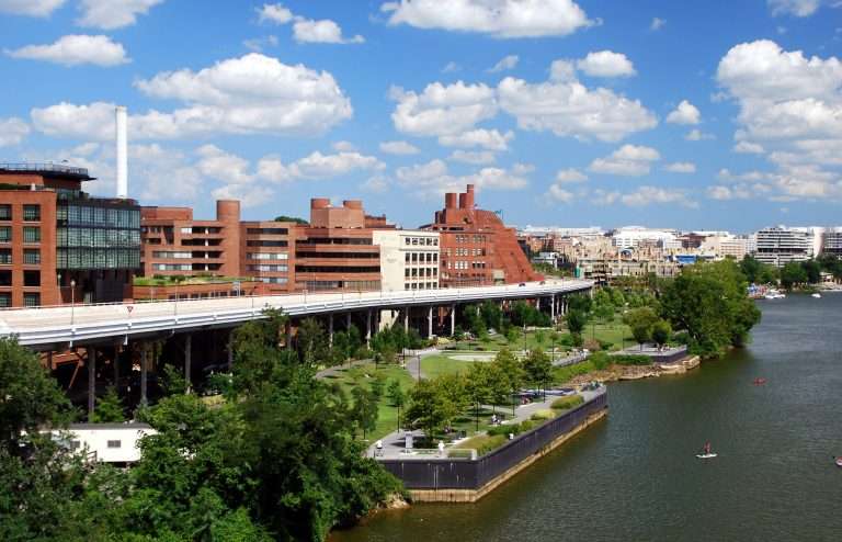 Aerial view of Georgetown Waterfront Park in Washington, D.C., featuring landscaped green spaces along the Potomac River, red-brick buildings, walking paths, and people kayaking and paddleboarding on a sunny day with blue skies and scattered clouds.