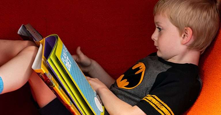 A young boy in a Batman shirt reclines against a bright red background while reading a large hardcover book titled "Plant-Based Diet." His legs are bent, and he is deeply focused on the pages, embodying a quiet moment of concentration and learning at the Hyattsville Library.