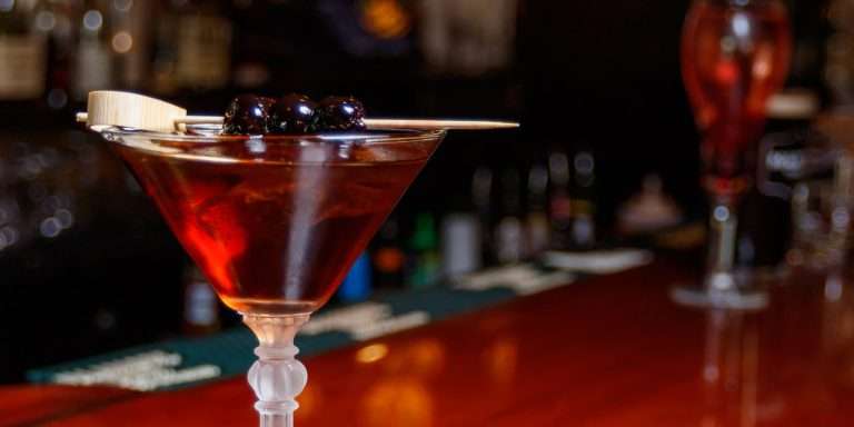 Elegant dark amber cocktail in martini glass garnished with cherries and lemon twist on polished bar surface with blurred restaurant background at Martin's Tavern Georgetown