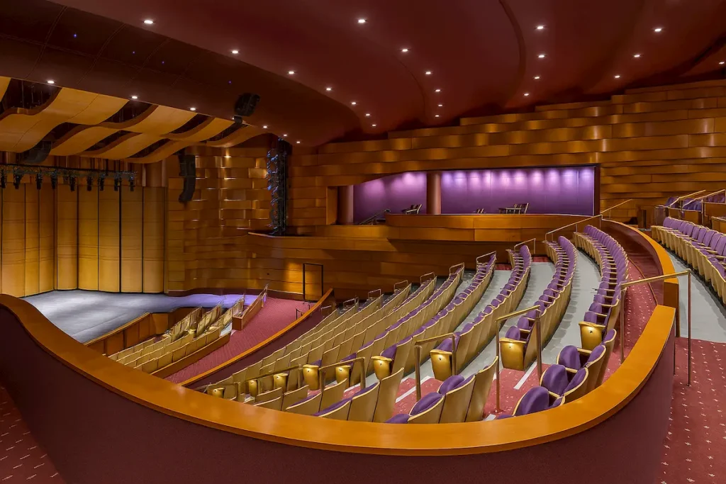 A modern theater interior featuring a large stage, curved rows of tan seats with purple cushions, a balcony level, and warm wooden and maroon architectural details, all under recessed ceiling lighting.