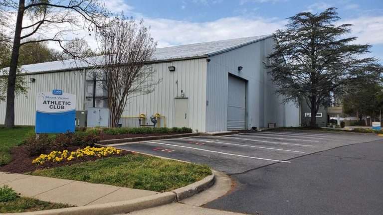 Mount Vernon Athletic Club exterior - gray metal building with blue and white signage, parking lot, and landscaped entrance with yellow flowers under cloudy sky