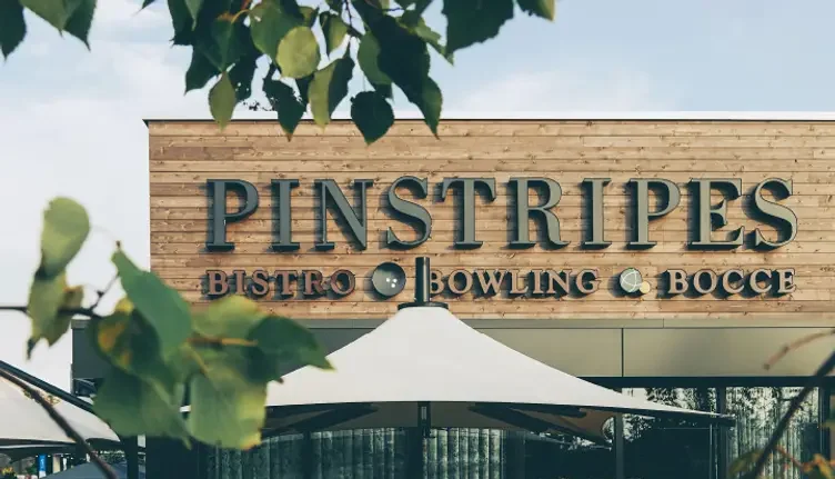 Guests enjoying food and drinks on the patio of Pinstripes Bistro, surrounded by flowers and shaded by large umbrellas, with the building’s wooden façade and “Pinstripes Bistro Bowling Bocce” signage visible through leafy branches.