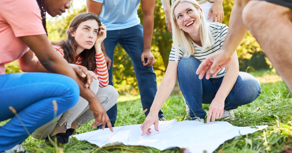 A diverse group of adults crouches in a park, examining a blank map or paper on the grass as they strategize during a scavenger hunt.
