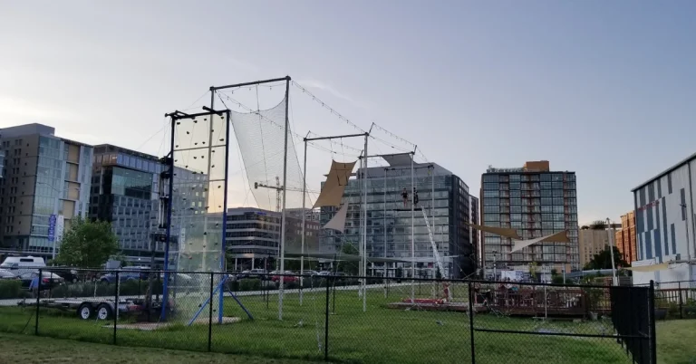 View of trapeze rig and climbing wall setup at Navy Yard in Washington, D.C., with high-rise buildings in the background and people enjoying aerial activities.