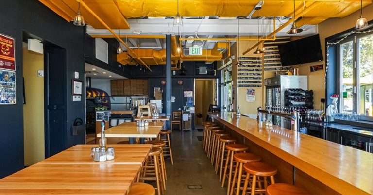 A modern pizzeria interior featuring a long wooden bar with matching stools, wooden tables, and a cozy, industrial design with exposed yellow ceiling ducts, hanging pendant lights, a beer tap wall, and a casual dining atmosphere.