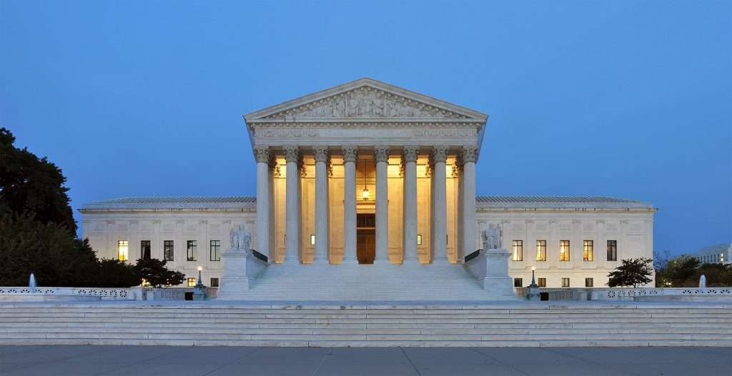 United States Supreme Court building. The neoclassical structure is illuminated against a twilight sky. It features a prominent facade with a series of large columns supporting a pediment. The inscription "EQUAL JUSTICE UNDER LAW" is visible above the columns. Stairs lead up to the entrance, with statues flanking the steps.