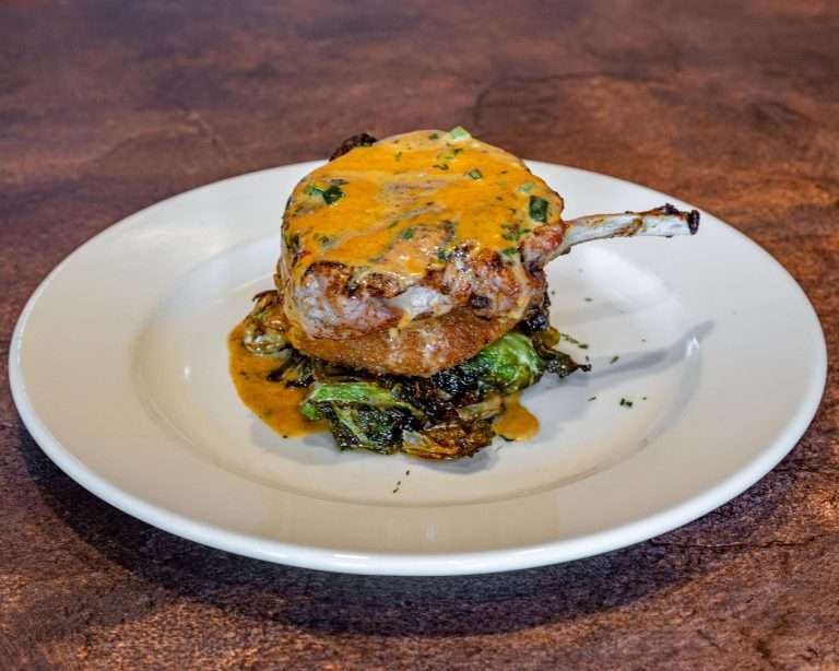 The Light Horse restaurant gourmet pork chop topped with melted cheese and herbs, served over sautéed Brussels sprouts with white sauce drizzle on elegant white plate