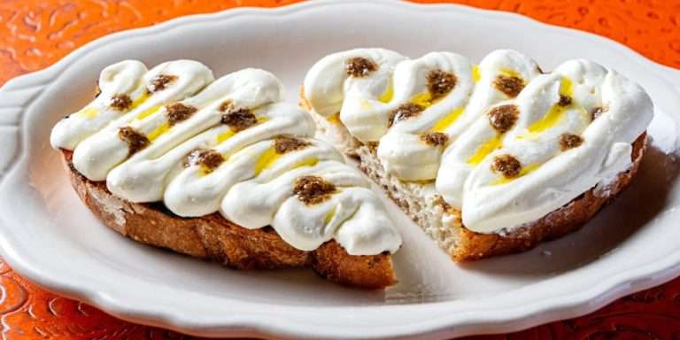 Artisanal toast topped with creamy white spread, golden olive oil drizzle, and brown seasoning dots on white ceramic plate at The Red Hen Georgetown Italian restaurant