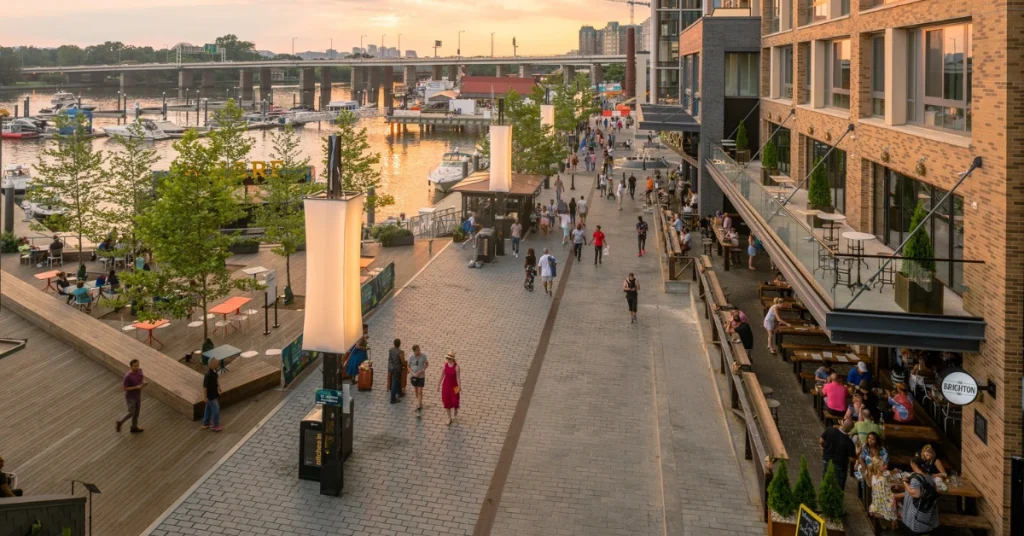 A bustling scene at The Wharf Promenade with people strolling, shops lining the walkway, boats docked in the harbor, and clear blue sky overhead.