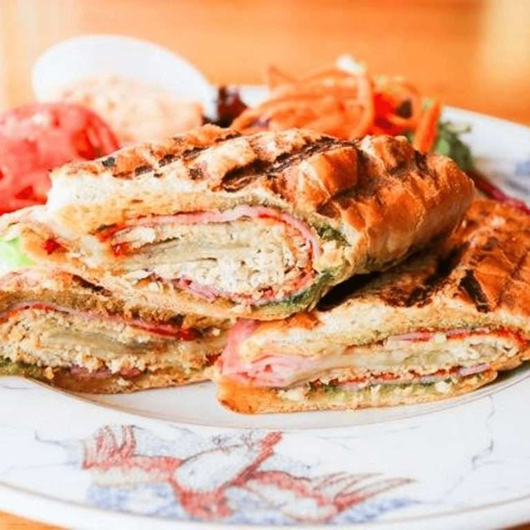A grilled sandwich stacked with layers of meat, cheese, and vegetables, served with a side of fresh tomato and salad on a decorative plate at Cleveland Park Bar & Grill.