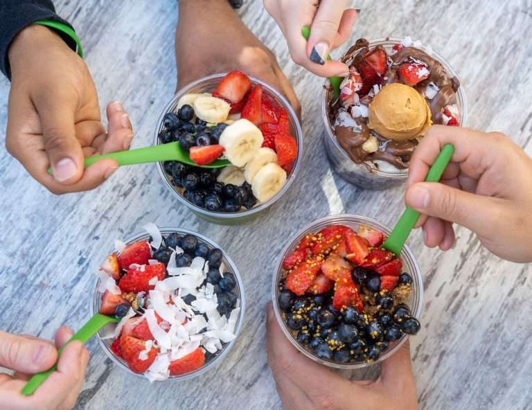 Four hands holding vegan-friendly acai bowls from South Block topped with fresh strawberries, blueberries, banana slices, coconut flakes, and various healthy toppings with green spoons
