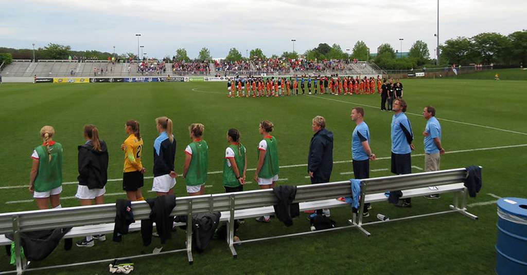 Washington Spirit players and coaching staff line up on the sideline before an NWSL match, with both teams lined up on the field and fans in the stands in the background.