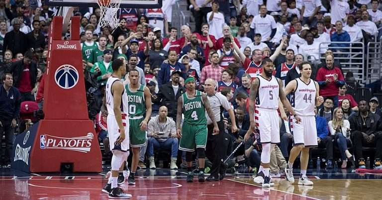 Washington Wizards players face off against Boston Celtics players during an NBA playoff game, with a packed arena of fans in the background cheering and watching intensely.