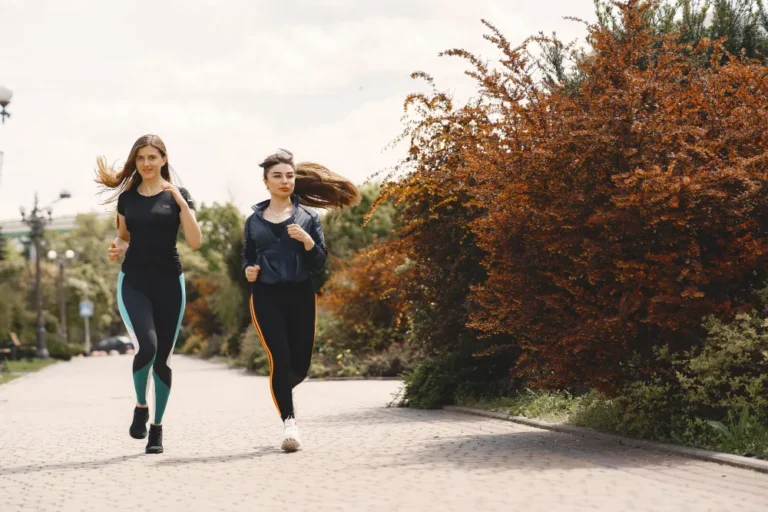 Two young women jogging outdoors on a paved path, surrounded by greenery and autumn-colored bushes.