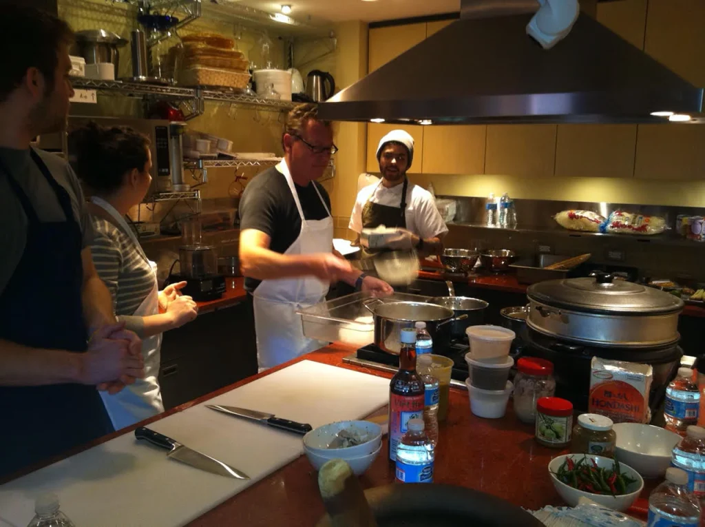 This appears to be a cooking class or a private cooking demonstration in a home kitchen. Several people are participating, with one chef-like figure (the man in the white apron) seemingly leading the instruction. The others are observing and assisting. They are preparing some kind of dish, possibly Asian cuisine, given the ingredients visible (like rice noodles and what looks like chili peppers). The kitchen is well-equipped with both standard appliances and specialized cooking tools like a steamer basket. The atmosphere seems relaxed and informal.