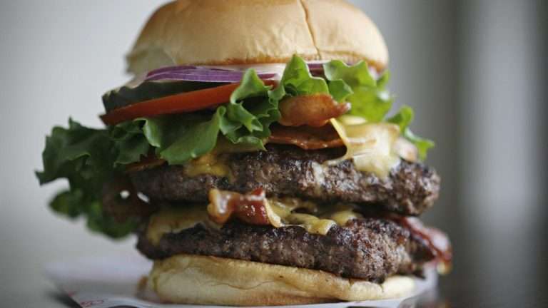 Double cheeseburger loaded with lettuce, tomato, onion, bacon, and cheese on a bun from Good Stuff Eatery, showcasing American cuisine.