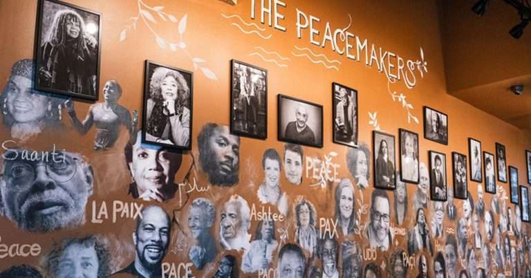 A vibrant mural featuring painted portraits of influential figures, titled "The Peacemakers," with their images surrounded by words for “peace” in various languages. Black-and-white framed photographs of the individuals hang above the painted faces, set against an orange wall.