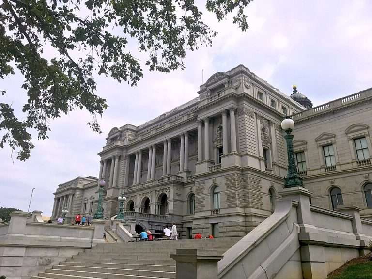 Library of Congress in Washington, D.C. The building has a grand staircase leading up to it, with people sitting and walking on the steps. There are trees and vintage-style lampposts around the area, contributing to the stately atmosphere of the scene.