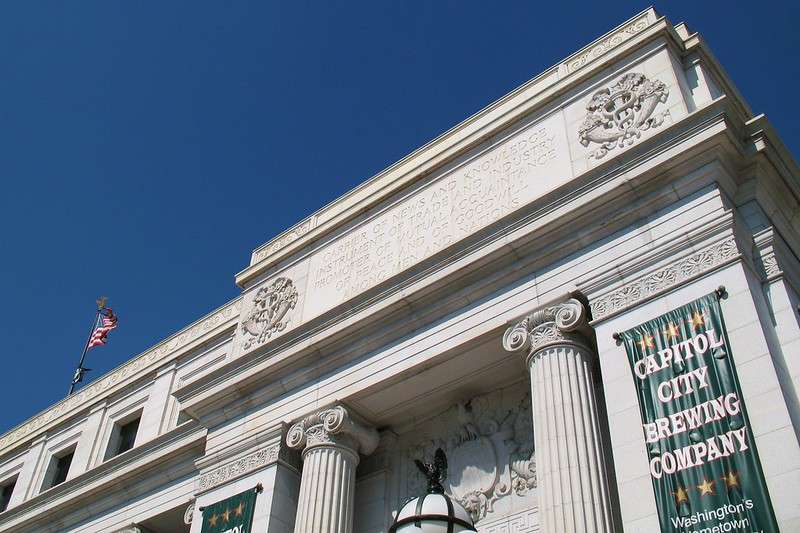 Low angle view of a portion of a white marble building with columns, decorative carvings, an American flag, and a "Capitol City Brewing Company" banner.