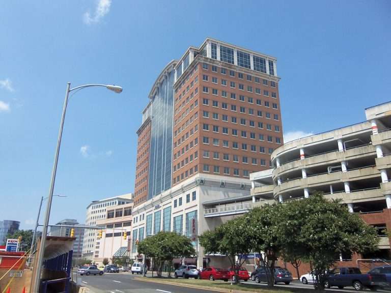 Tall red-brick building with glass windows, adjacent to a circular parking structure under a clear blue sky.