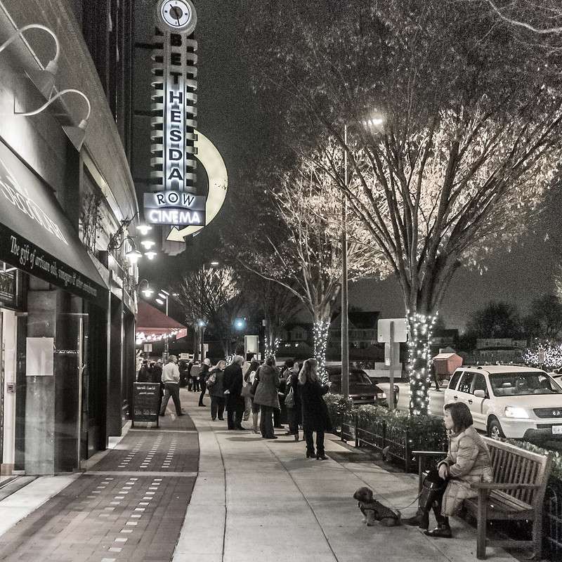 Nighttime city sidewalk with a line of people outside a cinema.