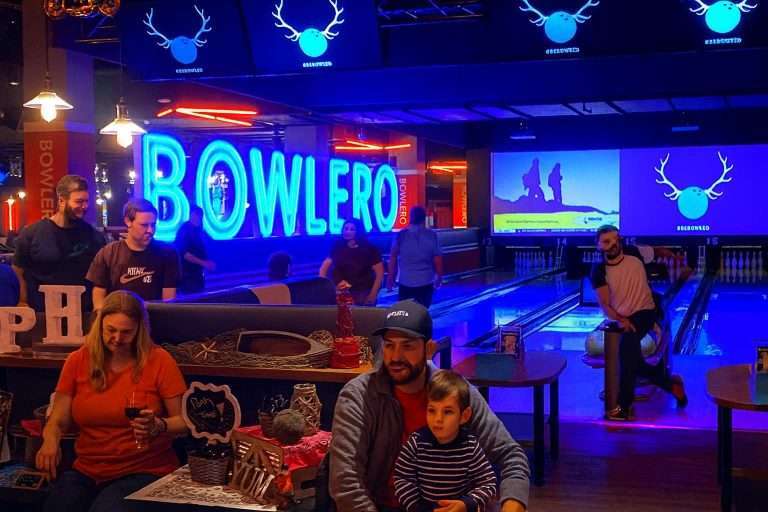 People bowling in a vibrant alley with neon "BOWLERO" sign, screens, and party decorations; a man and child in foreground.