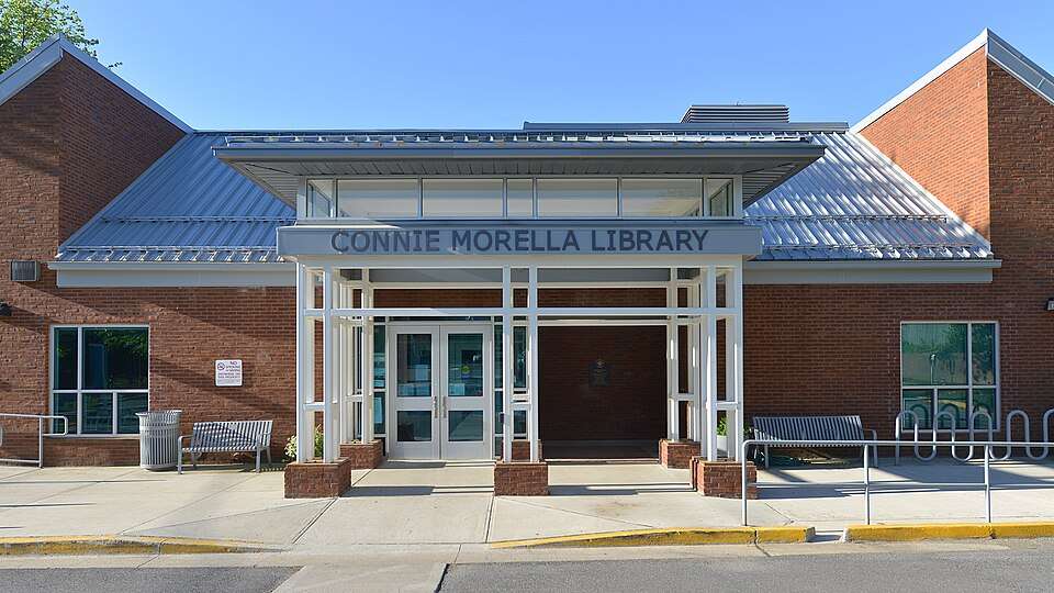 Brick library entrance with metal roof, white pillars, benches, and bike racks in front; labeled "Connie Morella Library".