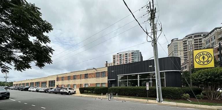 Street view of a Gold's Gym with a large sign, adjacent to a brick building and parked cars, under a cloudy sky.