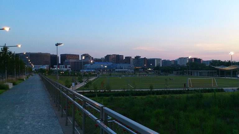Pathway and soccer field with goalposts under evening sky, city skyline in background.