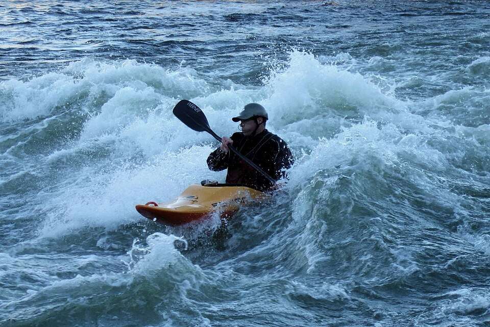 Kayaker navigating through rough water with turbulent waves, wearing a cap and holding a paddle.