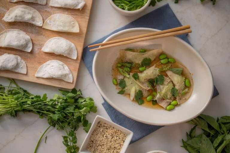 Dumplings in a bowl with broth, garnished with edamame and herbs, alongside uncooked dumplings on a board.