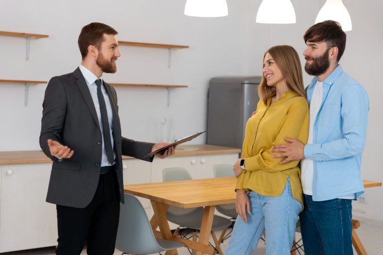 Three people in a modern kitchen, one in a suit holding a clipboard, and a couple smiling and listening attentively.