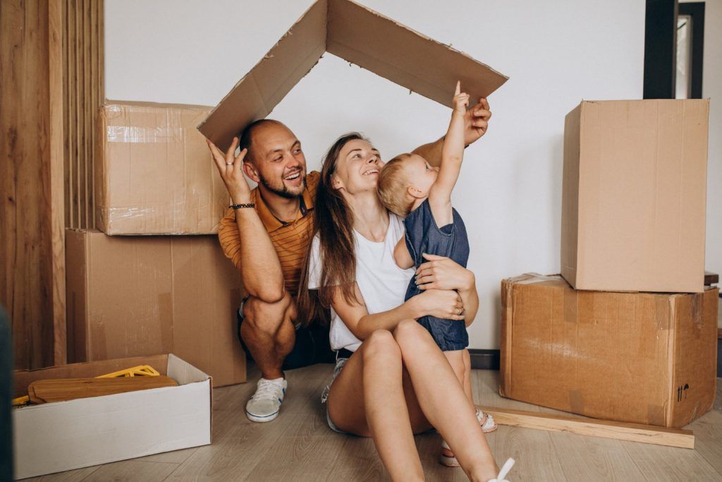 Family sitting among moving boxes, smiling, with a cardboard roof overhead.
