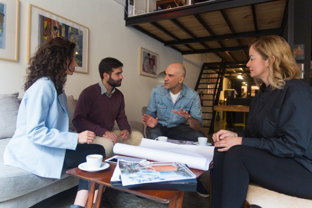 Four people in discussion, seated around a table with papers and coffee cups, in a cozy, art-adorned room.